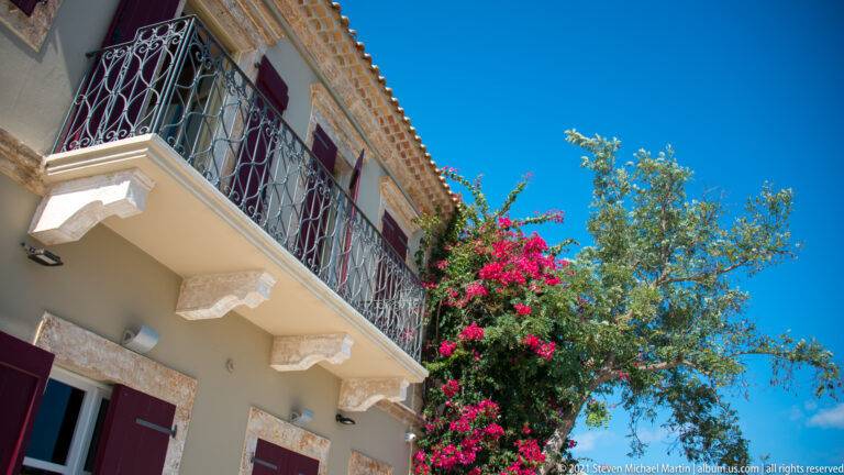 Upward view of an ornate iron balcony beside bright bougainvillea on a pale Fiskardo house.