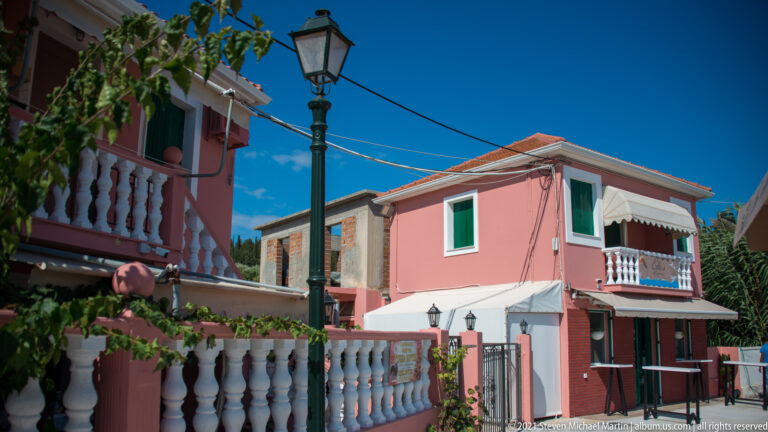 Black street lamp between pink houses with white balconies, green shutters, vines, and overhead wires in Fiskardo.
