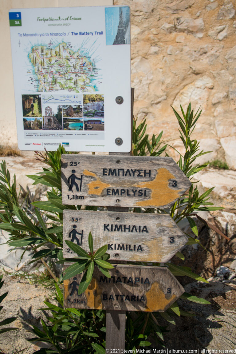 Weathered wooden walking-trail arrows with Greek and English place names beside a trail information board.