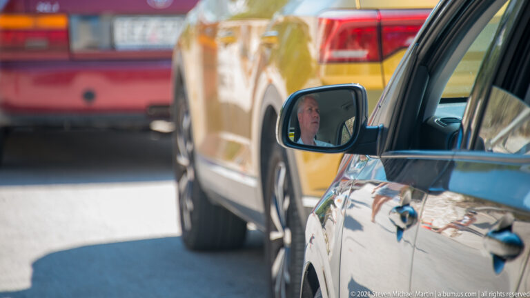 Driver reflected in a car side mirror with red, yellow, and white vehicles queued closely behind.