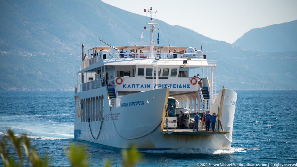 White vehicle-and-passenger ferry seen from the stern with open car deck and mountainous coast behind.