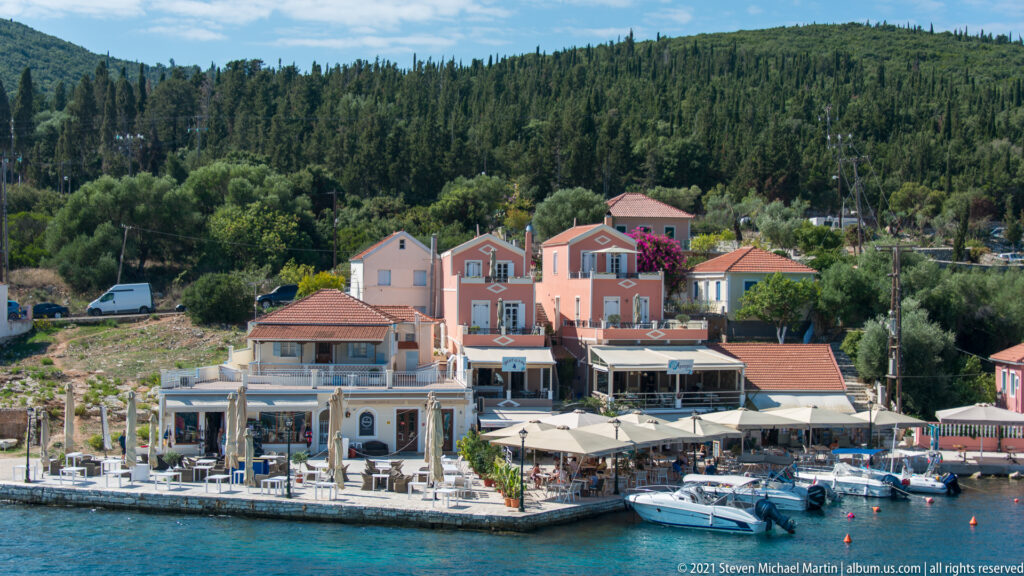 Pastel waterfront buildings, restaurant umbrellas, small boats, and a wooded hillside along Fiskardo harbor.