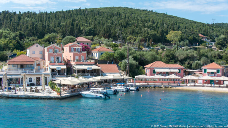 Wide view across turquoise water to Fiskardo’s pastel waterfront, moored boats, restaurant awnings, and wooded ridge.