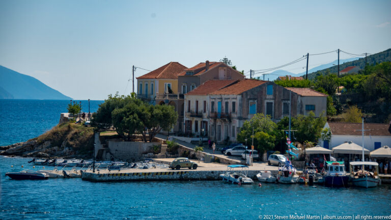 Weathered waterfront buildings with red-tiled roofs beside a small quay and moored boats in Fiskardo.
