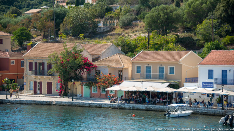 Pastel shuttered houses and a restaurant terrace under white umbrellas beside Fiskardo’s harbor.