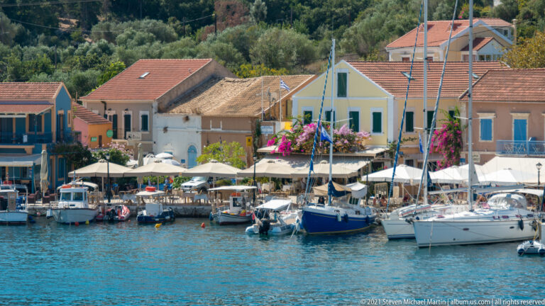 Sailing yachts moored closely before colorful Fiskardo buildings and restaurant terraces with white canopies.