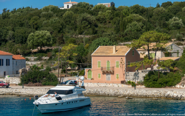 White cabin motor yacht on blue water before a peach house, stone seawall, and wooded hillside.