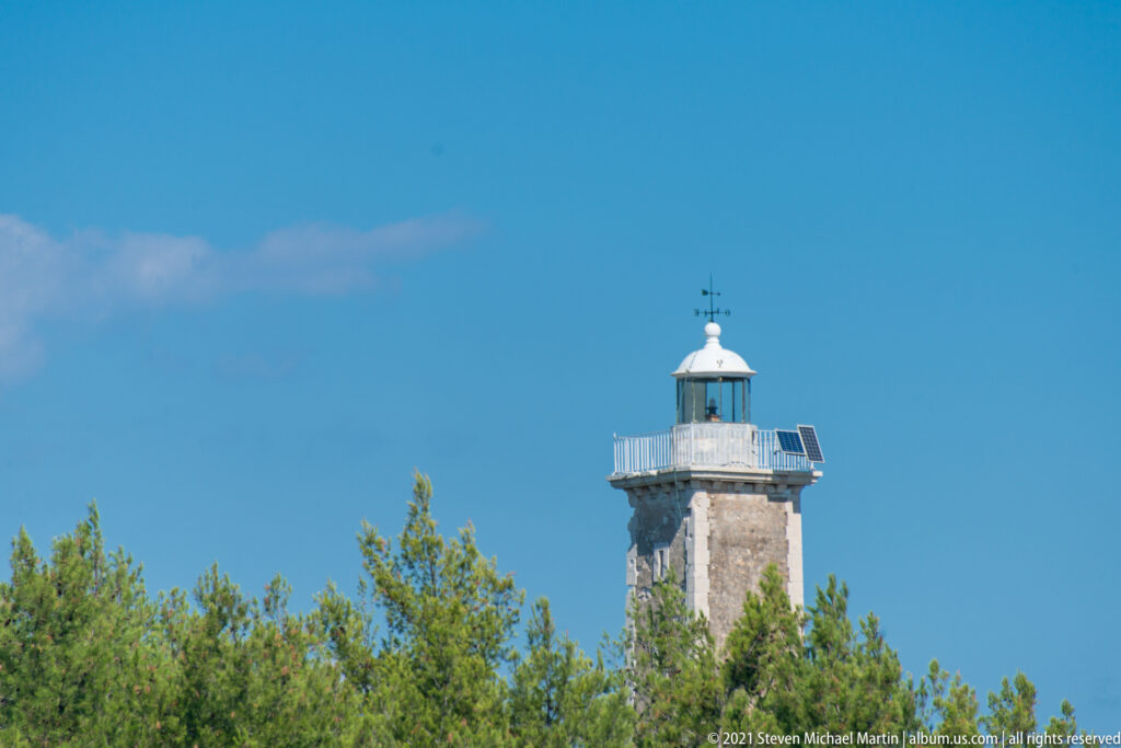 Square stone lighthouse tower with lantern gallery rising above green pine trees under a clear blue sky.