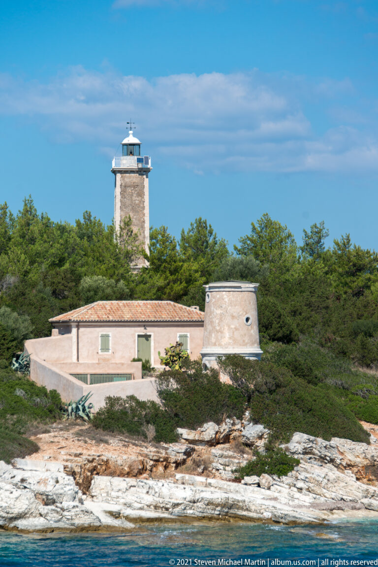Stone lighthouse tower behind a low keeper’s building and tapered round masonry structure on a rocky coast.