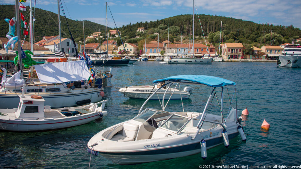 White motorboat with blue canopy among small boats and yachts, with Fiskardo’s waterfront buildings across the harbor.