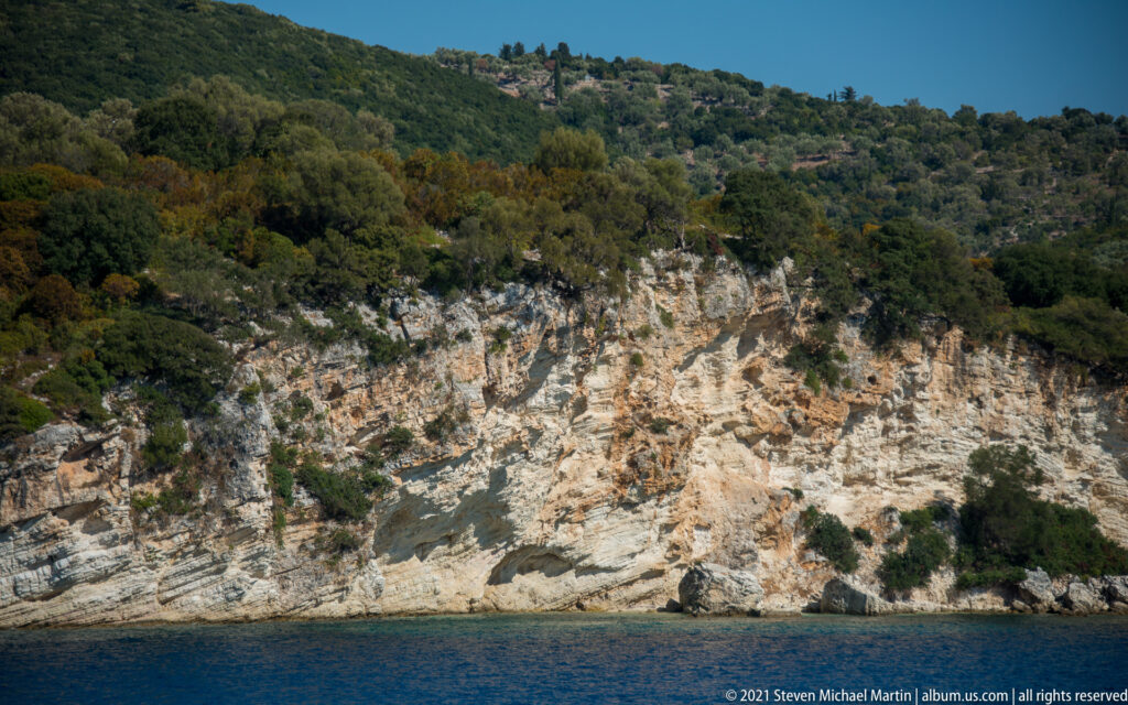 Pale layered rocky cliff dropping into deep blue water beneath a densely wooded hillside near Fiskardo.
