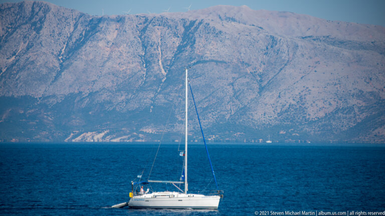White single-mast cruising sailboat on deep blue water with a massive gray mountain rising across the channel.