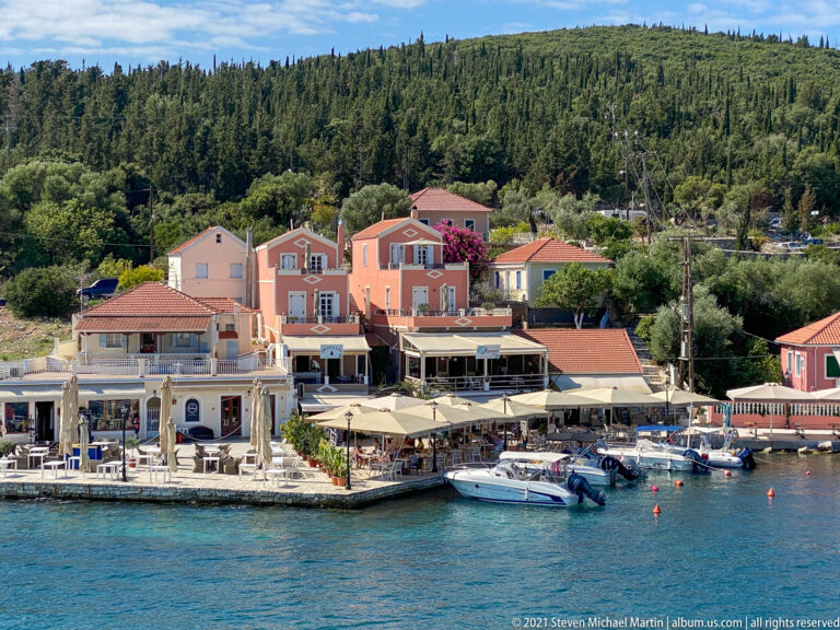 Peach and pink waterfront buildings, restaurant umbrellas, and small boats along Fiskardo harbor beneath a wooded hill.
