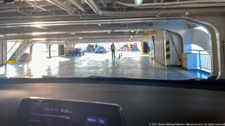 Ferry vehicle deck seen over a car dashboard, with steel columns, overhead beams, vehicles, and people ahead.