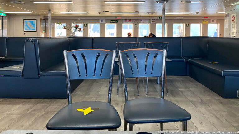 Dark bench seating and metal chairs with yellow spacing markers inside a bright passenger ferry lounge.