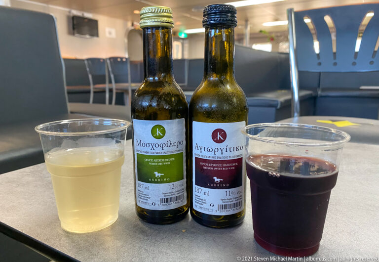 Small bottles labeled Moschofilero and Agiorgitiko beside plastic cups of white and red wine on a ferry table.