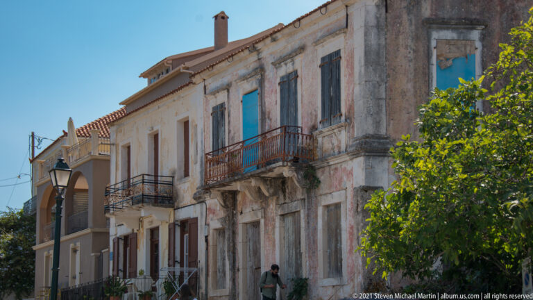 Weathered two-story masonry façade with tall shutters, iron balconies, faded plaster, and a red-tiled roof.