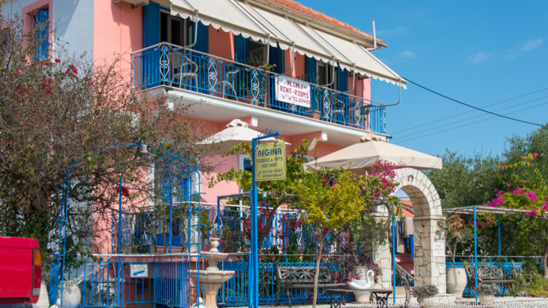 Pink building with blue balcony railings, striped awnings, flowering plants, and accommodation signs near the entrance.