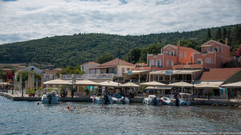 Waterfront cafés under awnings beside moored boats, with a salmon-colored building and wooded hillside behind.