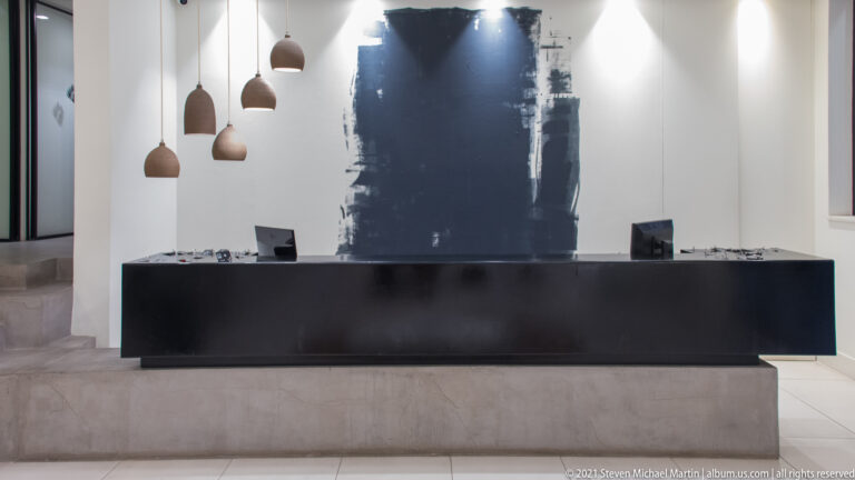 Long black reception desk beneath pendant lights, backed by a large dark abstract brushstroke on a white wall.