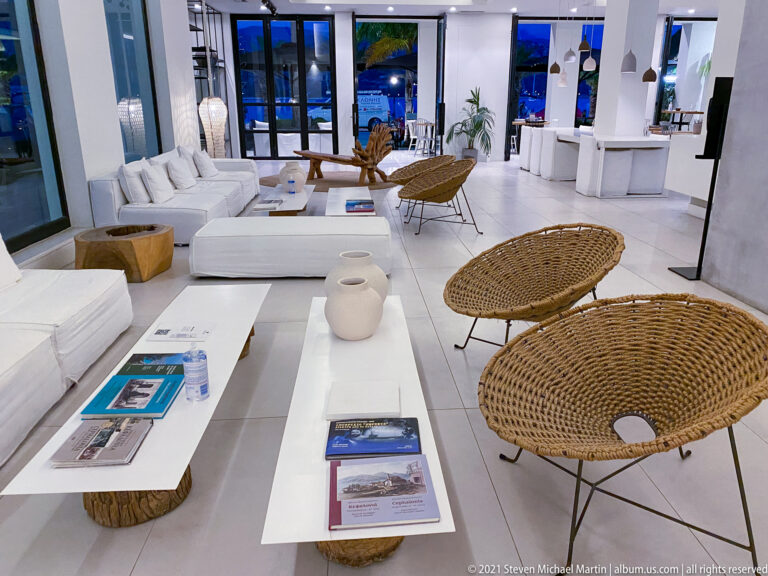 Bright white hotel lounge with sofas, low tables, woven chairs, books, and windows opening toward blue evening light.
