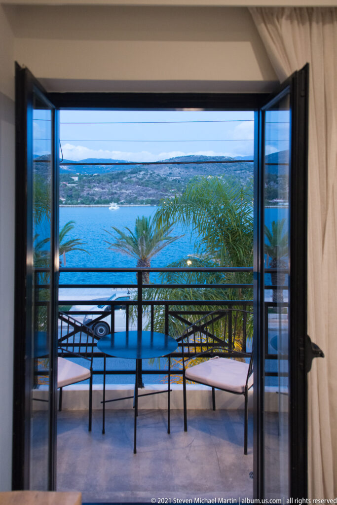 Open doorway framing a balcony bench, palm fronds, blue bay, and low shoreline in the distance.
