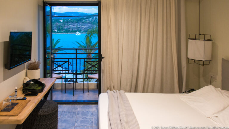 Compact hotel room with white bed, desk, wall-mounted television, and balcony overlooking bright blue water and green shore.
