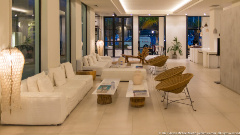 Warmly lit white lounge with long sofas, woven armchairs, low tables, and glass doors at the far end.