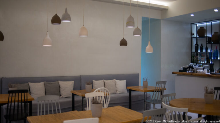 Small wooden tables and chairs beside a gray banquette and compact bar, with pendant lights overhead.