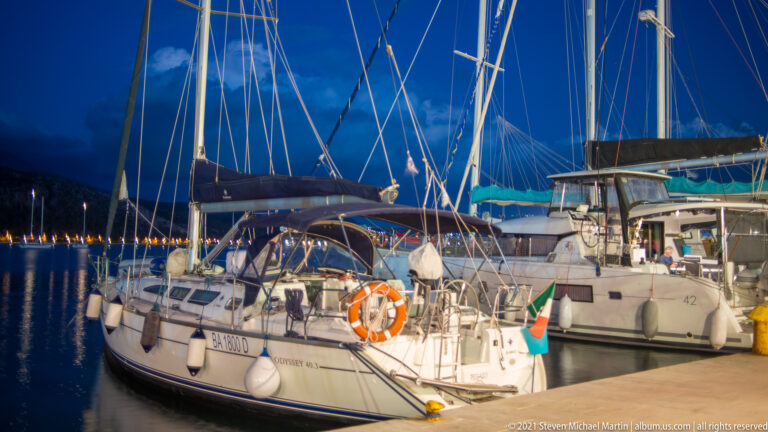 Moored sailboats seen from the stern at night, with tall masts, rigging, fenders, life ring, and harbor reflections.