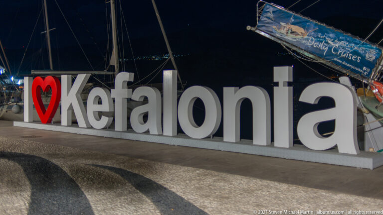 Illuminated white Kefalonia letters and red heart at night, with marina masts and signage behind.
