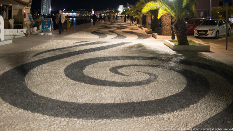 Black-and-white spiral pebble paving illuminated at night along a palm-lined waterfront promenade.