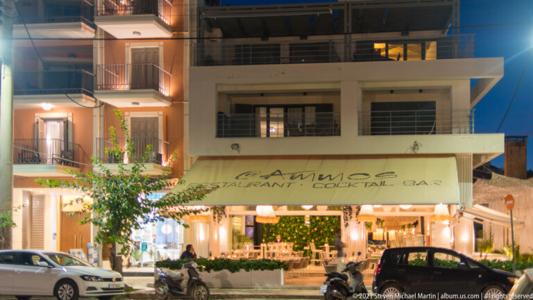 Illuminated MAMMA Restaurant Cocktail Bar façade at night, with diners, awning, balconies, scooters, and parked cars.