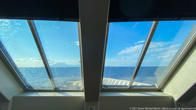 Blue sea and distant mountainous coastline viewed through large sloping windows inside a ferry or passenger vessel.