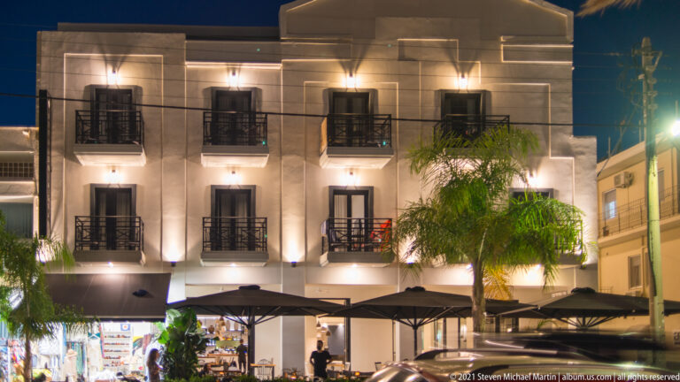 Warmly illuminated pale building façade with dark balconies, decorative wall bands, palm trees, and café umbrellas at street level.