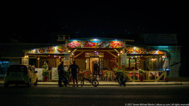 Bright roadside restaurant at night with food photographs across the fascia, people at the entrance, parked vehicles, and bicycle.