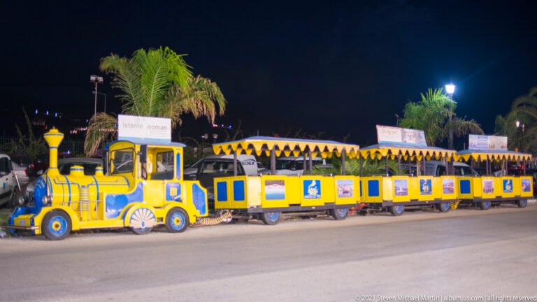 Yellow-and-blue tourist road train with locomotive-style front and several open passenger carriages parked beneath palm trees at night.