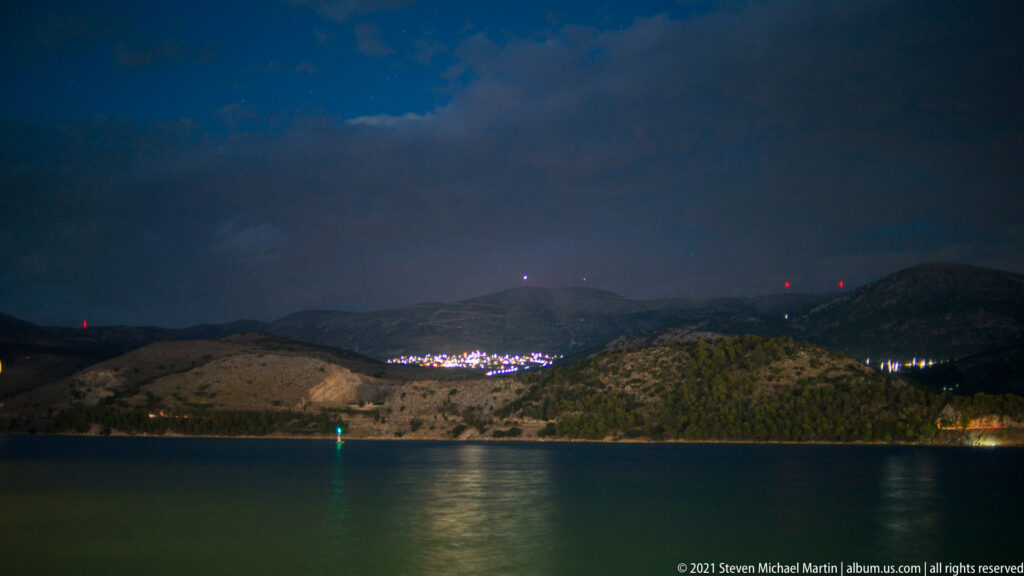 Dark coastal bay beneath hills at night, with shoreline lights, red hilltop beacons, and a green navigation light reflected on water.