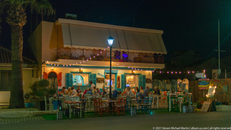 Busy open-air restaurant at night with diners at colorful tables beneath an awning, balcony, string lights, and streetlamp.
