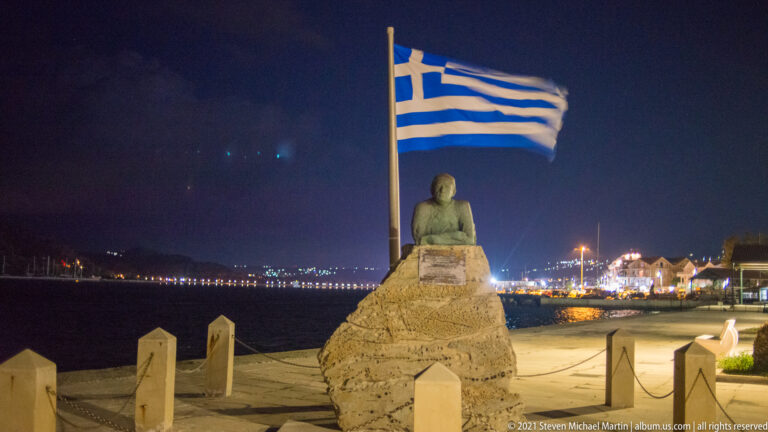 Seated figure monument on a rough stone pedestal beneath a large Greek flag, with harbor lights across the water at night.