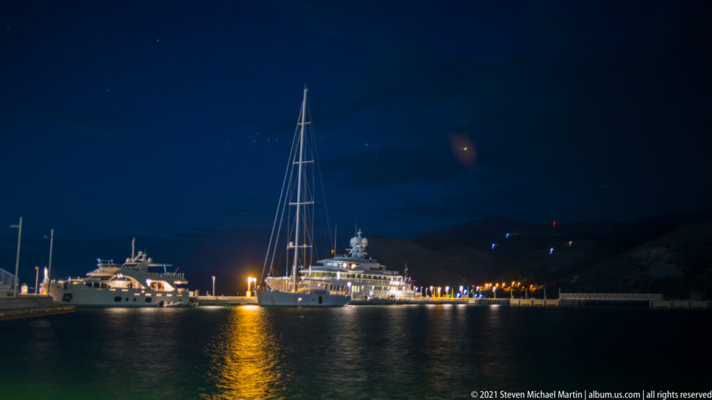 Large sailing yacht and motor yachts moored across dark harbor water, with golden lights reflecting toward the camera.