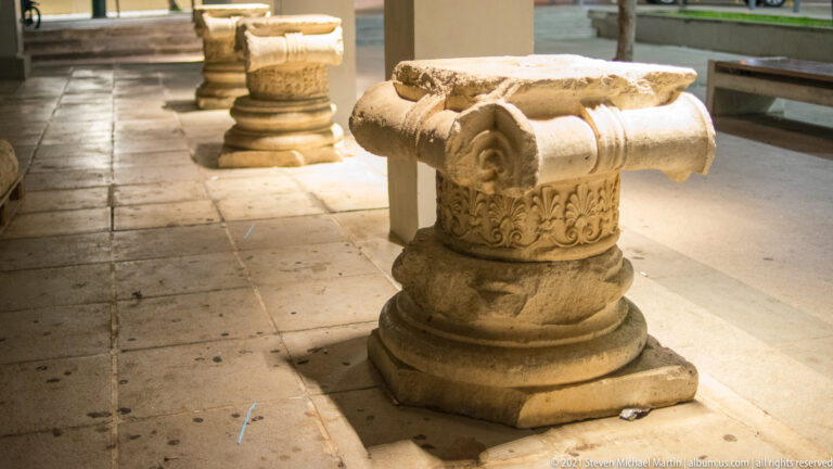 Weathered carved stone capital with volute-like corner scrolls and vegetal ornament displayed on a low pedestal.