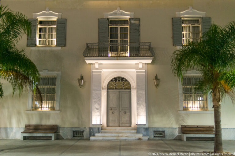 Symmetrical pale façade at night with central arched doorway, small balcony, shuttered windows, benches, and palm trees.