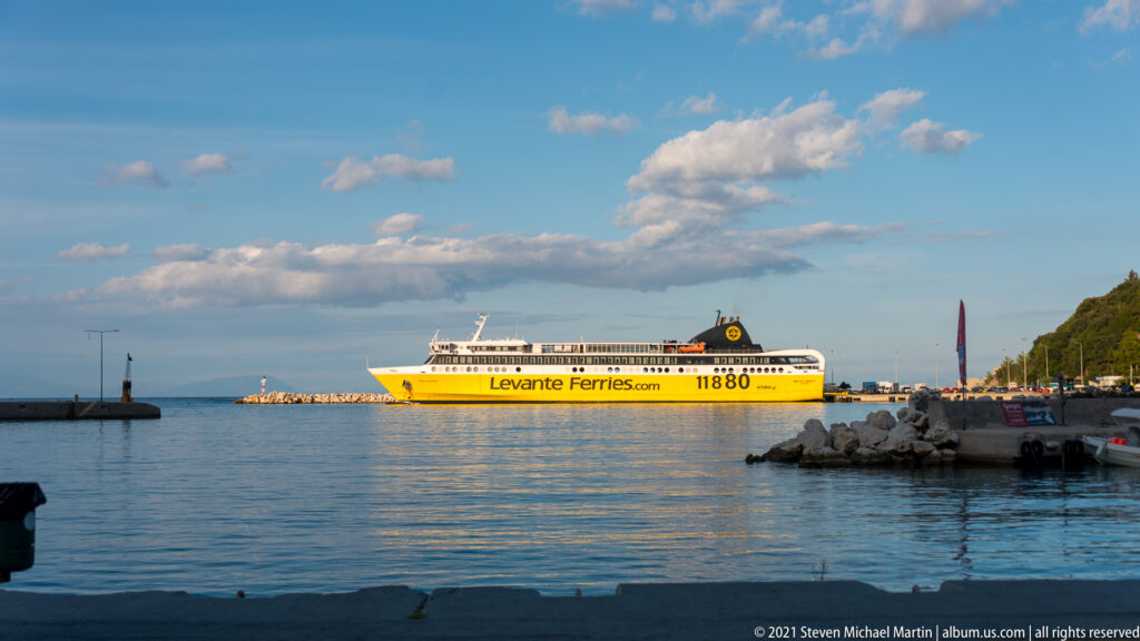 Yellow-and-white Levante Ferries vessel crossing calm harbor water, with breakwaters, small boats, and wooded shoreline beyond.