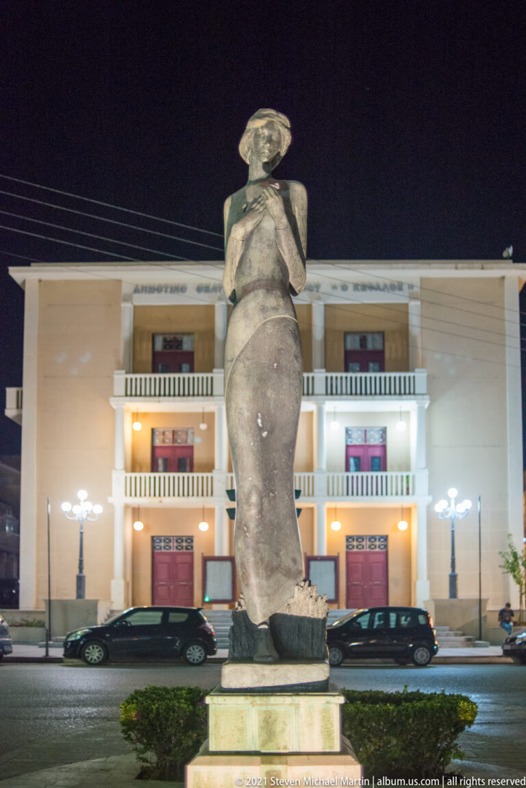 Tall abstract standing human figure on a stone plinth at night, with an illuminated multi-story building behind.