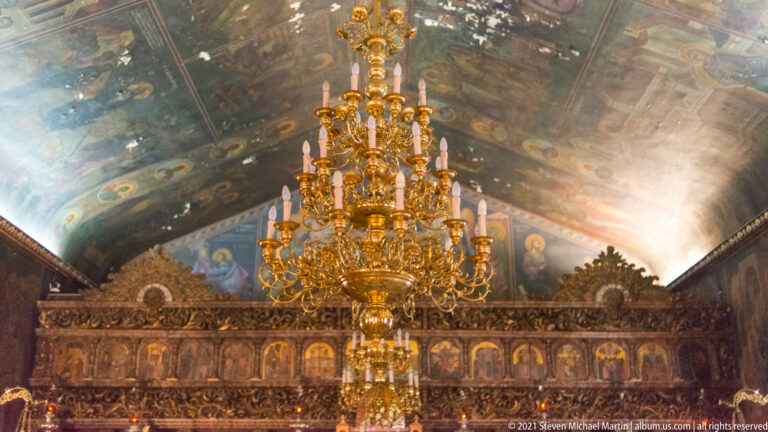 Large gilded chandelier hanging before an ornate iconostasis beneath a richly painted vaulted Orthodox church ceiling.