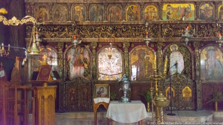 Ornately carved Orthodox iconostasis with painted icons, hanging lamps, candlestands, and a small table in front.