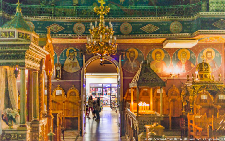 Orthodox church nave with painted saints, chandeliers, carved wooden furnishings, candles, and an open central doorway.