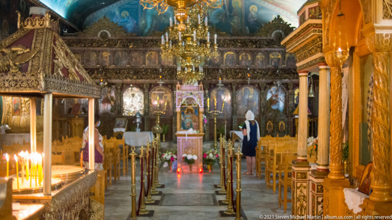 Central aisle of an Orthodox church leading to a carved iconostasis, with chandelier, candles, chairs, and one visitor.