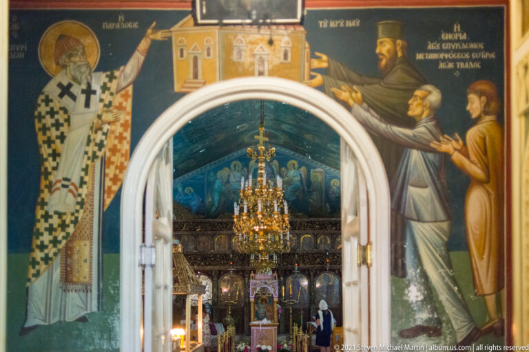 Painted clerical figures surround a white arched doorway opening into an Orthodox church nave and iconostasis.